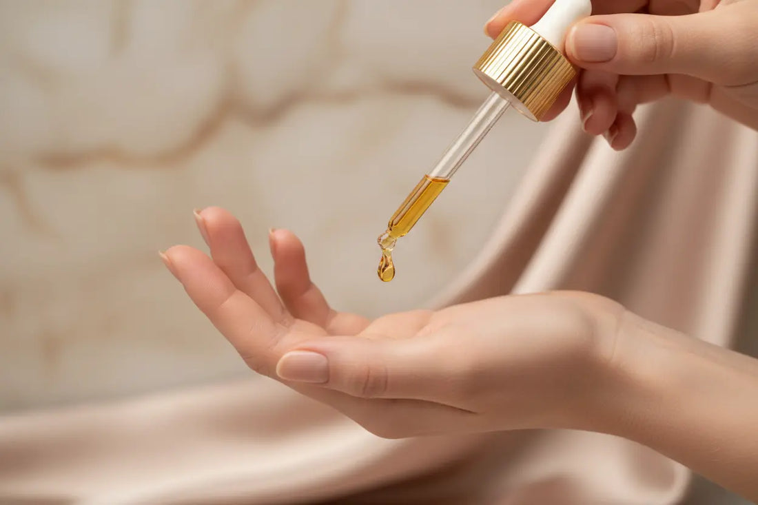 Droplet of golden perfume oil falling from a dropper onto a hand, set against a marble and soft fabric background.